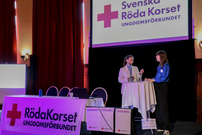 Generalsekreterare Beatrice Amsenius och Förbundsordförande Lotta Schneider står på en scen med Röda Korsets Ungdomsförbunds logotyp på en skärm i bakgrunden. 